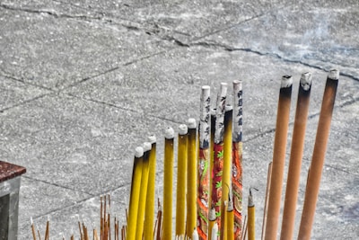 A collection of incense sticks in varying sizes and colors, with some featuring decorative patterns and characters. The sticks are arranged in a holder on a concrete surface, and a few of them are partially burned, emitting faint smoke.