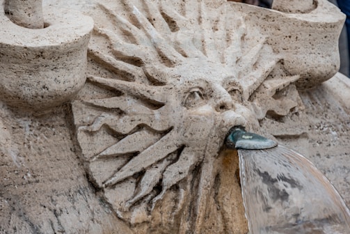 A detailed stone carving portrays a face with flowing hair or rays around it, through which water is emerging from its mouth. The stone appears aged and textured, contributing to an ancient or classical aesthetic.
