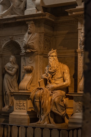 A detailed sculpture depicting a seated bearded figure, intricately carved from marble. The setting includes ornate architectural elements and additional sculptures, highlighting an ornate, classical style. The lighting accentuates the textures and details of the sculpture.