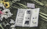 An open notebook on grass, surrounded by white and yellow flowers. The notebook contains a handwritten message expressing sadness over someone's passing, with artistic decorations on the page.