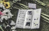 An open notebook on grass, surrounded by white and yellow flowers. The notebook contains a handwritten message expressing sadness over someone's passing, with artistic decorations on the page.