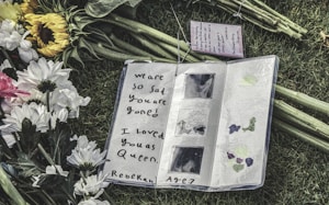 An open notebook on grass, surrounded by white and yellow flowers. The notebook contains a handwritten message expressing sadness over someone's passing, with artistic decorations on the page.
