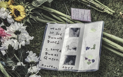 An open notebook on grass, surrounded by white and yellow flowers. The notebook contains a handwritten message expressing sadness over someone's passing, with artistic decorations on the page.