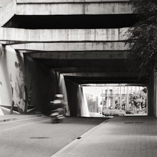 A black and white image depicting a motorcycle riding through an underpass. The underpass features concrete structures and a mural on one wall, partially visible in the shadows. Sunlight creates contrasting dark and bright areas. In the background, a modern building and some trees can be seen.
