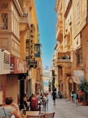 A narrow, sunlit street lined with historic stone buildings and shops, with signs in various languages. People are sitting at outdoor tables enjoying food or drinks, while others walk down the street. In the background, a glimpse of a distant body of water and a skyline is visible under a clear, blue sky.