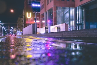 A nighttime urban scene with wet streets reflecting colorful neon lights. Buildings with glass facades and a prominent 'U' sign are visible, along with a pedestrian walkway. Streetlights line the road, casting a soft glow.
