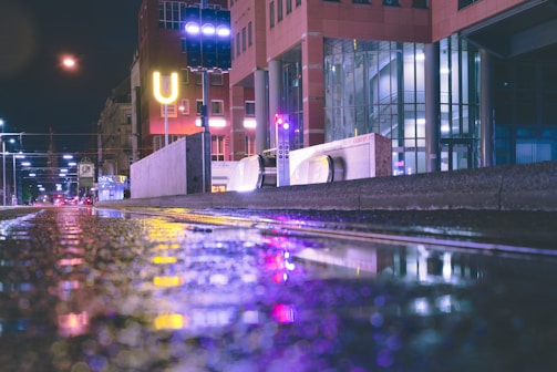 A nighttime urban scene with wet streets reflecting colorful neon lights. Buildings with glass facades and a prominent 'U' sign are visible, along with a pedestrian walkway. Streetlights line the road, casting a soft glow.