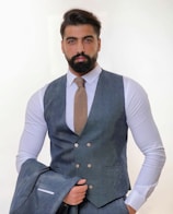 A man with a thick beard and neatly styled hair wears a sharply tailored gray suit with a matching waistcoat and a light brown textured tie. He holds the jacket of the suit in one hand while standing against a light, neutral background, giving an air of professionalism.