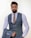 A man with a thick beard and neatly styled hair wears a sharply tailored gray suit with a matching waistcoat and a light brown textured tie. He holds the jacket of the suit in one hand while standing against a light, neutral background, giving an air of professionalism.