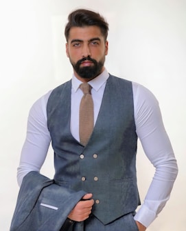 A man with a thick beard and neatly styled hair wears a sharply tailored gray suit with a matching waistcoat and a light brown textured tie. He holds the jacket of the suit in one hand while standing against a light, neutral background, giving an air of professionalism.