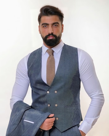 A man with a thick beard and neatly styled hair wears a sharply tailored gray suit with a matching waistcoat and a light brown textured tie. He holds the jacket of the suit in one hand while standing against a light, neutral background, giving an air of professionalism.