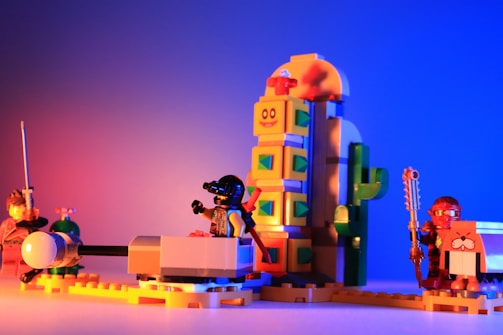 A vibrant assembly of LEGO characters arranged in a playful setup. On one side, a figure in armor wields a sword, while another in futuristic gear appears to be seated in a vehicle. A whimsical structure resembling a cactus with a smiley face towers in the background. The scene is set against a backdrop of rich gradients from red to blue.