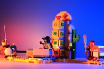 A vibrant assembly of LEGO characters arranged in a playful setup. On one side, a figure in armor wields a sword, while another in futuristic gear appears to be seated in a vehicle. A whimsical structure resembling a cactus with a smiley face towers in the background. The scene is set against a backdrop of rich gradients from red to blue.