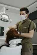 A dentist is attending to a patient in a dental clinic. The dentist is wearing a face mask and gloves, focusing on the task. The patient is seated in a dental chair with an overhead light, and there is a serene atmosphere.