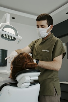 A dentist is attending to a patient in a dental clinic. The dentist is wearing a face mask and gloves, focusing on the task. The patient is seated in a dental chair with an overhead light, and there is a serene atmosphere.