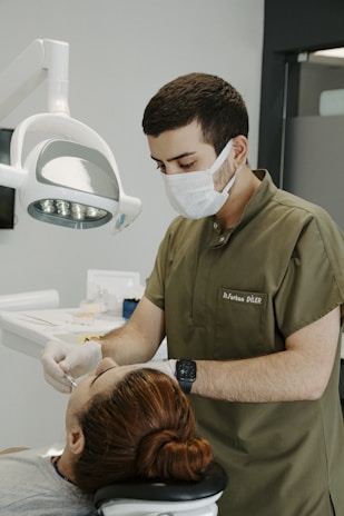 A dentist wearing a mask and green scrubs is attending to a patient lying in a dental chair. The dentist is focused on a procedure, using dental instruments under a bright overhead light. The patient has reddish-brown hair tied back and appears relaxed. Clinical tools and supplies are visible in the background, creating a professional and clean atmosphere.