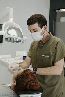 A dentist wearing a mask and green scrubs is attending to a patient lying in a dental chair. The dentist is focused on a procedure, using dental instruments under a bright overhead light. The patient has reddish-brown hair tied back and appears relaxed. Clinical tools and supplies are visible in the background, creating a professional and clean atmosphere.