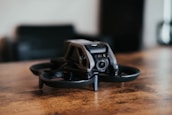 A small black drone with a sleek, modern design is placed on a wooden surface. The drone features protective guards around the propellers and a camera at the front. The background is blurred, suggesting an indoor setting.