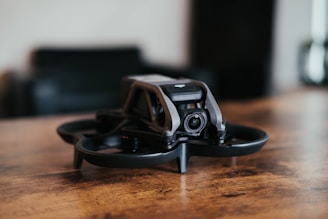 A small black drone with a sleek, modern design is placed on a wooden surface. The drone features protective guards around the propellers and a camera at the front. The background is blurred, suggesting an indoor setting.