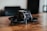 A small black drone with a sleek, modern design is placed on a wooden surface. The drone features protective guards around the propellers and a camera at the front. The background is blurred, suggesting an indoor setting.