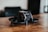 A small black drone with a sleek, modern design is placed on a wooden surface. The drone features protective guards around the propellers and a camera at the front. The background is blurred, suggesting an indoor setting.