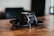 A small black drone with a sleek, modern design is placed on a wooden surface. The drone features protective guards around the propellers and a camera at the front. The background is blurred, suggesting an indoor setting.