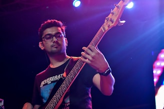 A person is playing an electric bass guitar on stage under colorful lighting. They are wearing glasses and a black t-shirt, with concentrated facial expression as they focus on their music.