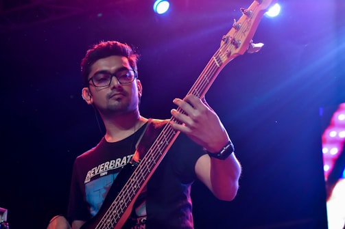 A person is playing an electric bass guitar on stage under colorful lighting. They are wearing glasses and a black t-shirt, with concentrated facial expression as they focus on their music.