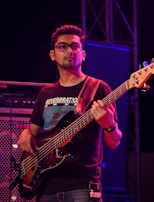 A musician is playing an electric bass guitar on stage. He is wearing glasses and a black t-shirt with text on it. The background includes speakers and stage lighting.