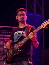 A musician is playing an electric bass guitar on stage. He is wearing glasses and a black t-shirt with text on it. The background includes speakers and stage lighting.