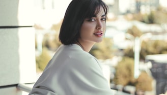 A woman with short dark hair gazes towards the camera while leaning against a railing. She wears a light-colored, flowing garment. The background is softly blurred, suggesting an outdoor urban setting.