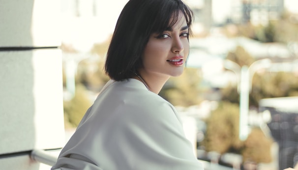 A woman with short dark hair gazes towards the camera while leaning against a railing. She wears a light-colored, flowing garment. The background is softly blurred, suggesting an outdoor urban setting.