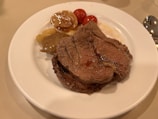 A plate with several pieces of cooked beef, accompanied by roasted tomatoes and caramelized onions. The beef appears juicy and well-cooked, with a slight sheen from the juices on top.