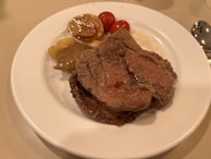A plate with several pieces of cooked beef, accompanied by roasted tomatoes and caramelized onions. The beef appears juicy and well-cooked, with a slight sheen from the juices on top.