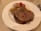 A plate with several pieces of cooked beef, accompanied by roasted tomatoes and caramelized onions. The beef appears juicy and well-cooked, with a slight sheen from the juices on top.