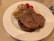 A plate with several pieces of cooked beef, accompanied by roasted tomatoes and caramelized onions. The beef appears juicy and well-cooked, with a slight sheen from the juices on top.
