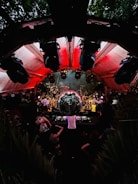 An energetic crowd gathers around a large disco ball in an outdoor venue with red lighting overhead. A DJ is performing at a console surrounded by people dancing and enjoying the lively atmosphere. The setting is surrounded by greenery and features various lights and speakers enhancing the party experience.