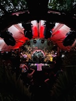 An energetic crowd gathers around a large disco ball in an outdoor venue with red lighting overhead. A DJ is performing at a console surrounded by people dancing and enjoying the lively atmosphere. The setting is surrounded by greenery and features various lights and speakers enhancing the party experience.