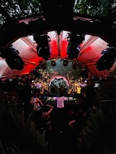 An energetic crowd gathers around a large disco ball in an outdoor venue with red lighting overhead. A DJ is performing at a console surrounded by people dancing and enjoying the lively atmosphere. The setting is surrounded by greenery and features various lights and speakers enhancing the party experience.