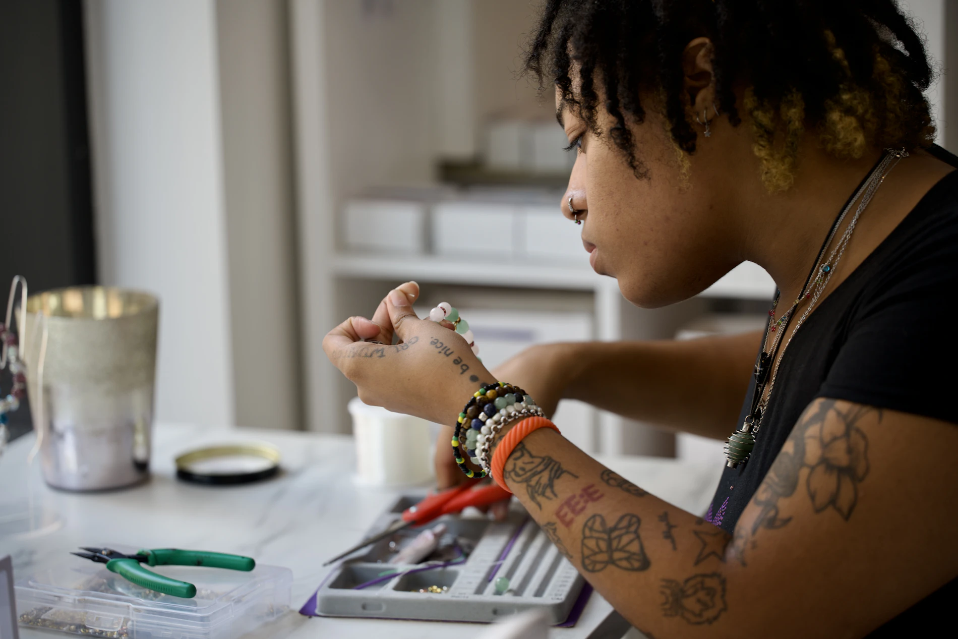 A jeweller leans into her bench, sorting stones from a tray with round-nose pliers, her own work stacked on her wrist.