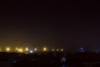 A night scene with an urban skyline featuring a series of streetlights casting a warm glow. A prominent arch structure is visible in the background, partially illuminated by the lights. The sky is enveloped in darkness, with minimal light pollution creating a tranquil atmosphere.