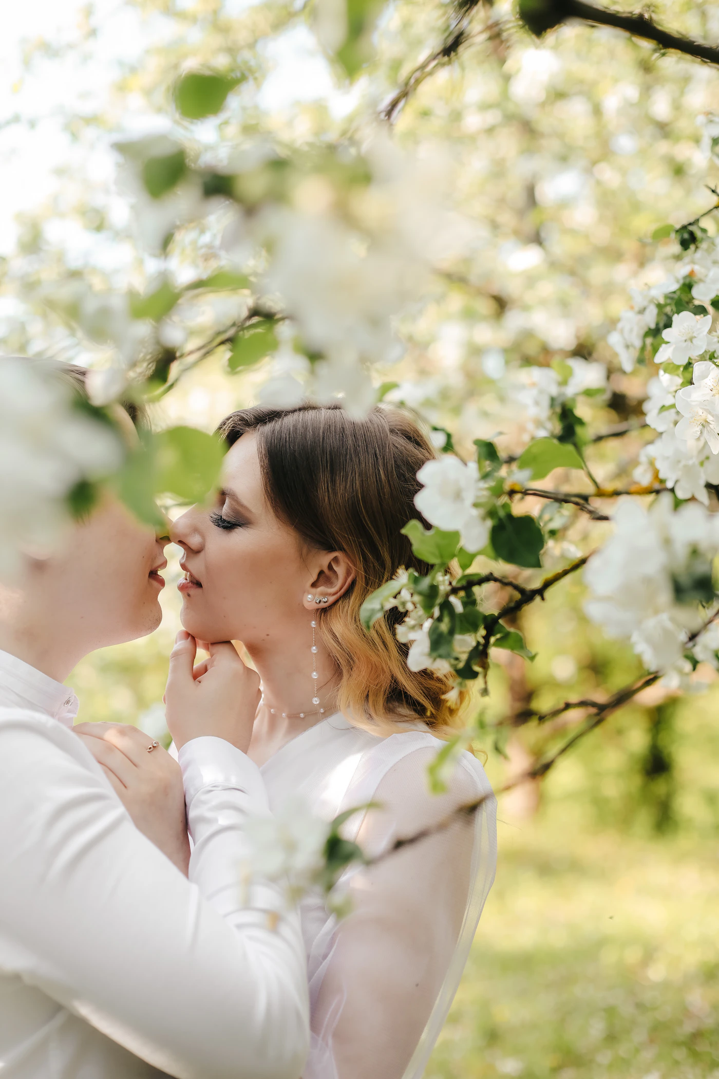 Couple in formal wedding attire sharing a tender kiss beneath the branches of a tree in soft light