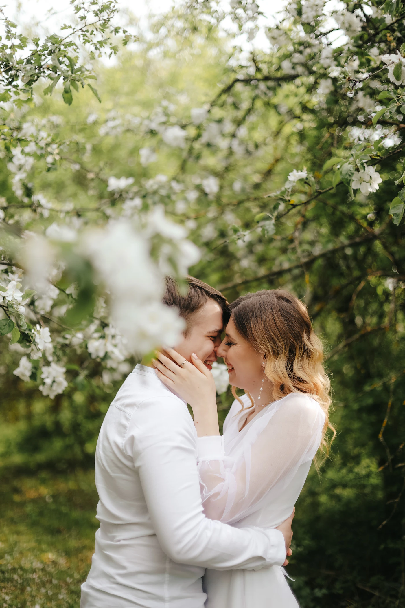 Couple in wedding attire sharing a kiss beneath the branches of a flowering tree in spring