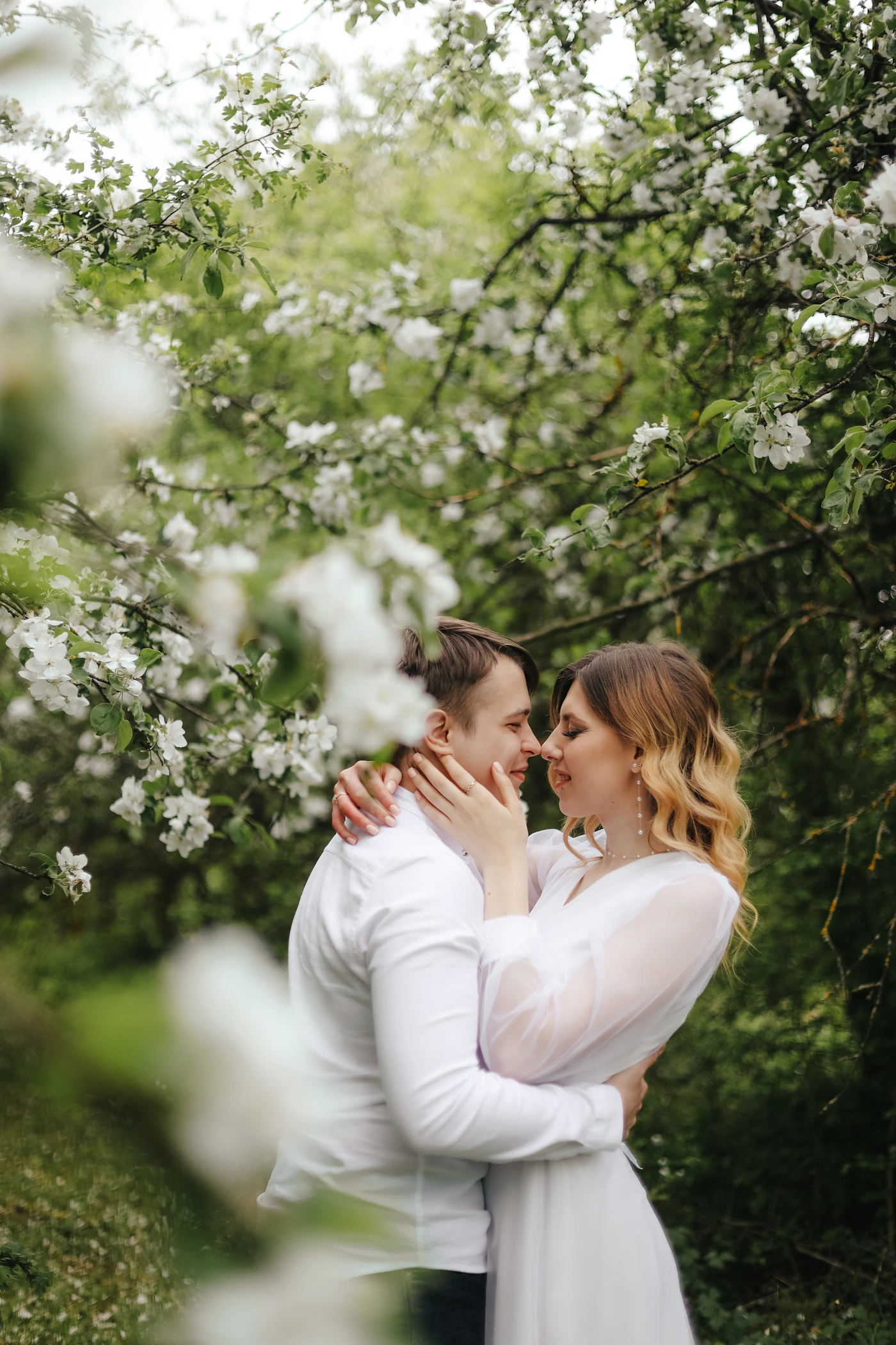 Bride in white gown kissing groom beneath a tree with soft natural light surrounding them