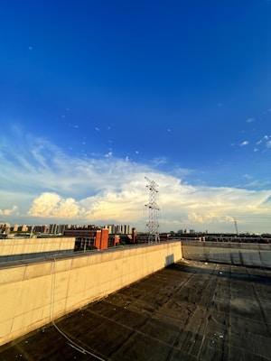 An expansive rooftop view overlooking a cityscape, featuring a vibrant blue sky with scattered clouds. A prominent power line tower stands tall near the horizon, surrounded by various buildings, including a red brick structure.