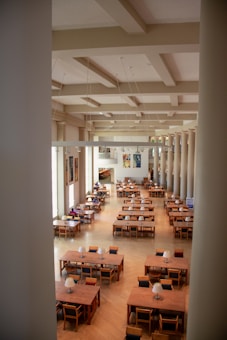 A spacious library or reading room with large wooden tables and chairs arranged neatly in rows. The room has high ceilings with large windows on one side, allowing natural light to flood in. Bookshelves and artwork line the walls, and there are a few people seated at the tables reading or working. The interior design features a classic style with pillars and subdued colors.