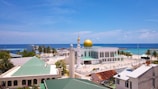 A coastal townscape featuring a prominent mosque with a large golden dome surrounded by several buildings with green-tiled roofs. Palm trees are scattered throughout, and the vivid blue ocean stretches out to the horizon under a clear sky.