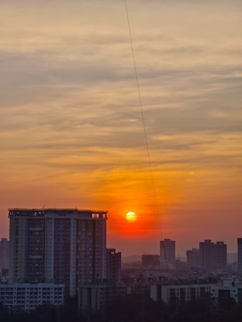 The image captures a city skyline during sunset. The sun is prominently visible, casting an orange and yellow hue across the sky, with some scattered clouds adding texture. Tall buildings form a silhouette against the vibrant sky, creating a picturesque urban landscape.