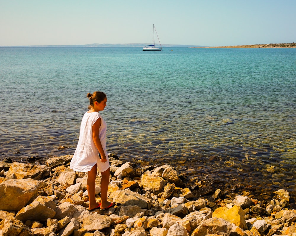 a girl standing on a rocky shore looking at a boat in the water