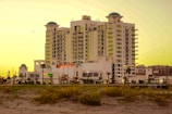 A multi-story hotel building with a white facade and several balconies, featuring rounded green domes on its roof. It is set against a golden sky, likely at sunset, with sandy foreground and green shrubs. Palm trees are visible near the entrance, and there is signage for a Subway restaurant on the ground floor.